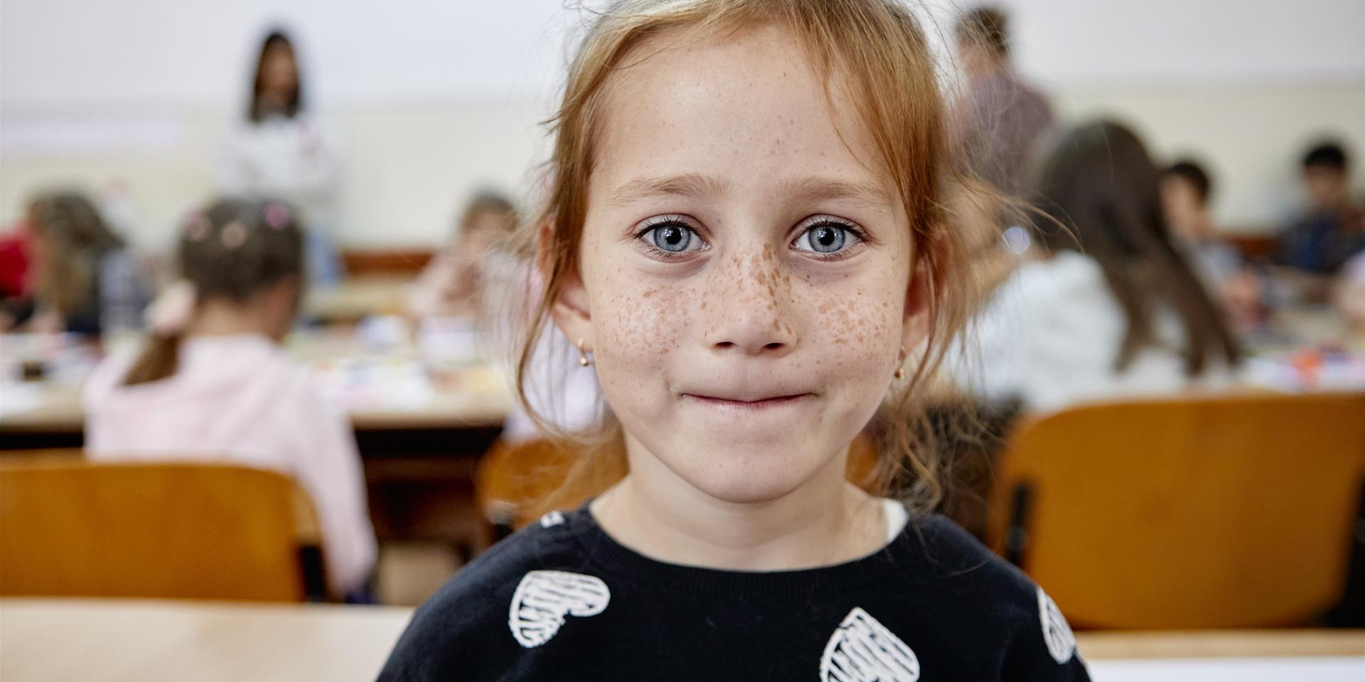 Una bambina ucraina sorride guardando l’obiettivo. Sullo sfondo una classe con banchi e bambini in aula in una scuola in Romania supportata da Save the Children