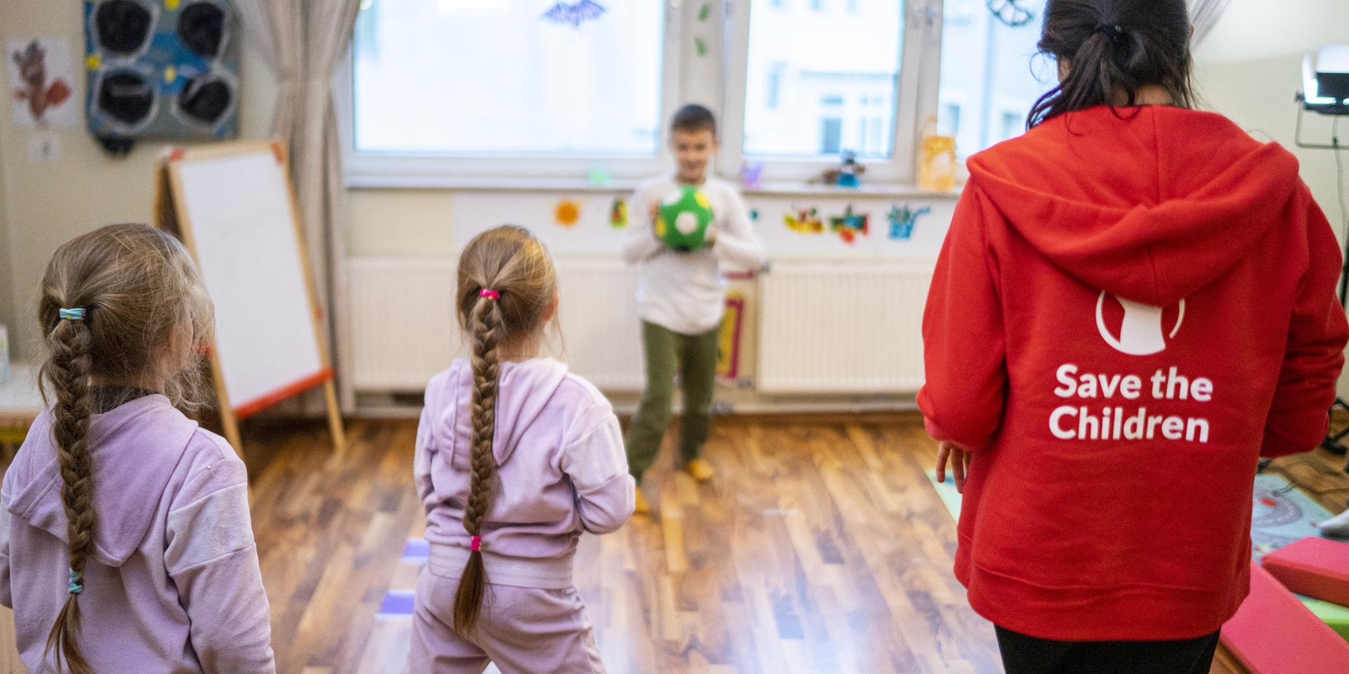 3 bambini ucraini giocano a palla sotto la supervisione di una operatrice Save the Children all’interno di uno Spazio a Misura di Bambino in Germania