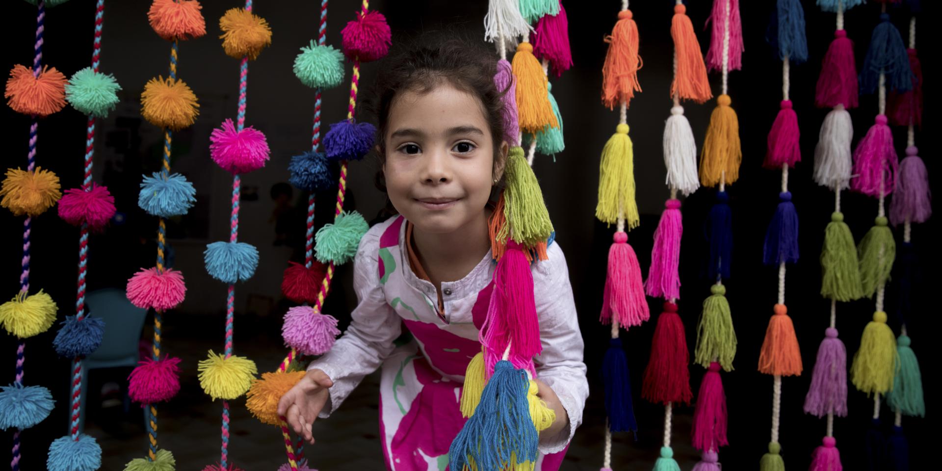 Una bambina di 5 anni spunta gioiosa da una tenda di pompom colorati in uno Spazio a Misura di Bambino per migranti venezuelani a Lima, in Perù.