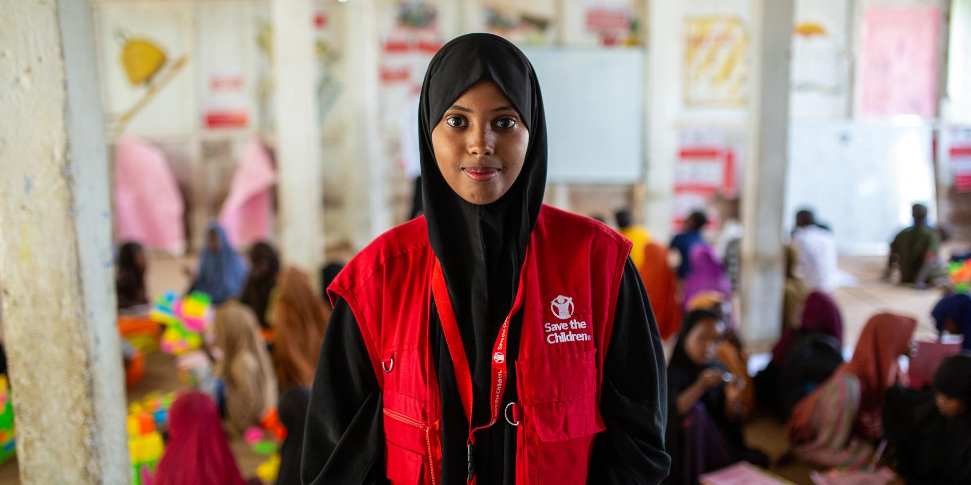 In primo piano un’operatrice guarda in camera con uno sguardo intenso. Sullo sfondo, i bambini e le bambine per cui opera all’interno di uno Spazio a Misura di Bambino di Save the Children in Somalia.