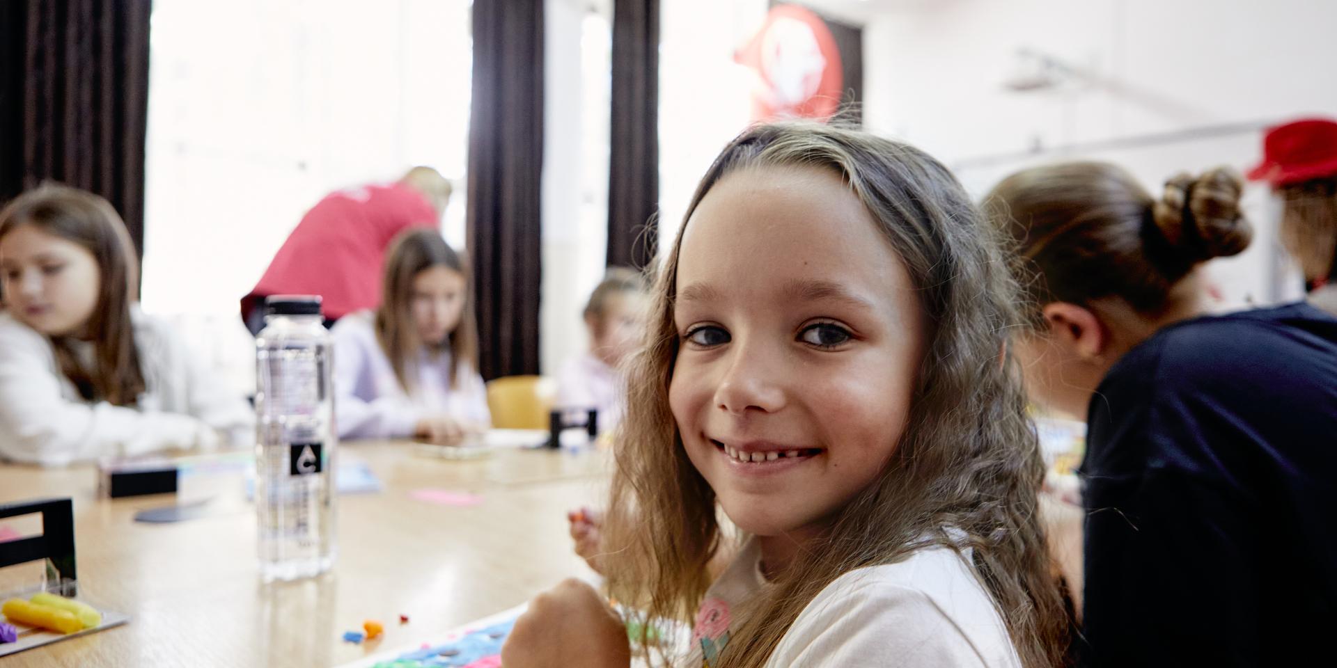 Una bambina sorride verso la camera mentre disegna sul banco con altre bambine all’interno di una scuola in Romania.