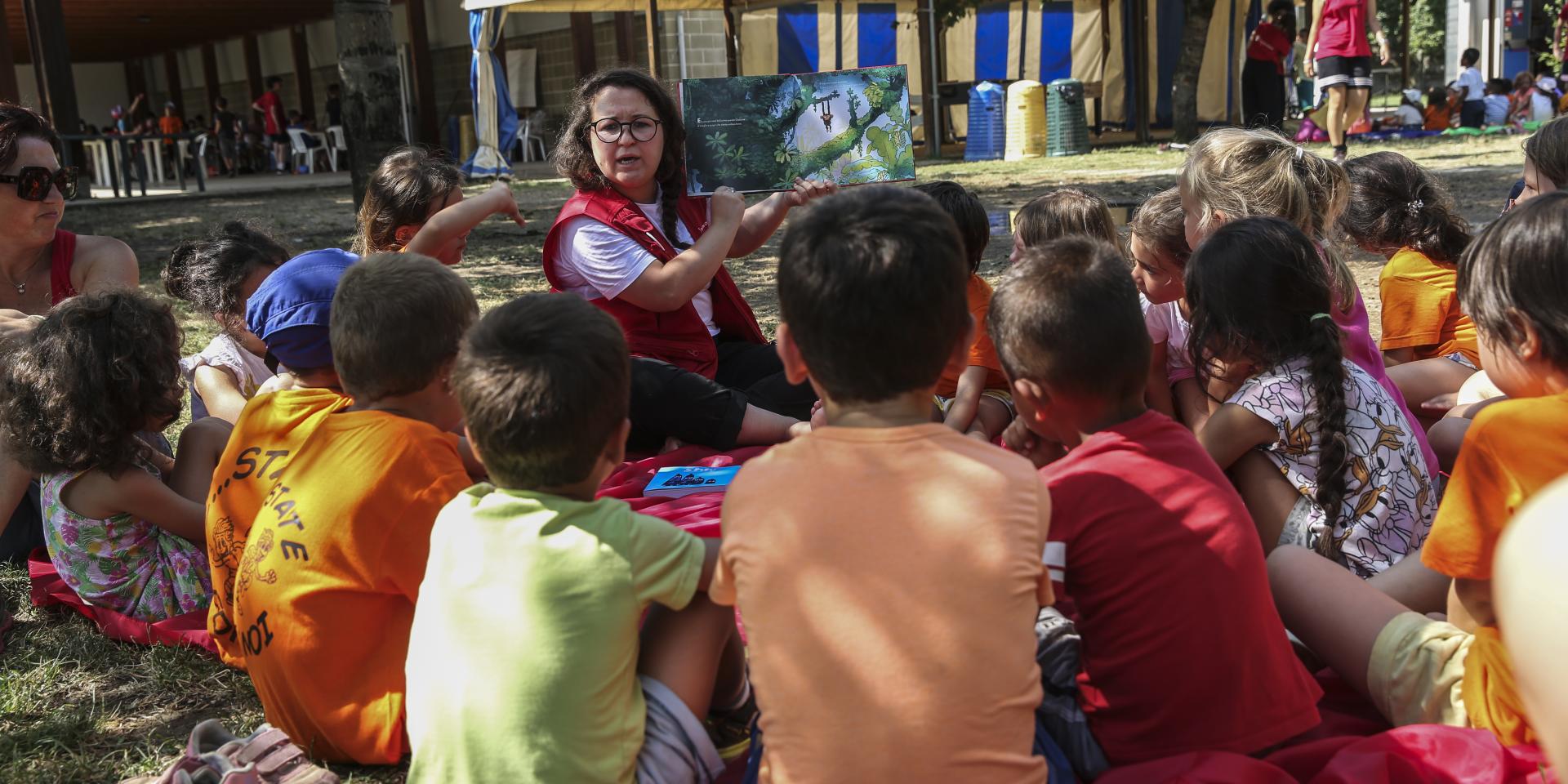 Un’operatrice legge un libro ad alta voce a bambine e bambini seduti in cerchio su un prato nell’ambito delle attività educative offerte nelle zone colpite dall’alluvione in Emilia Romagna.