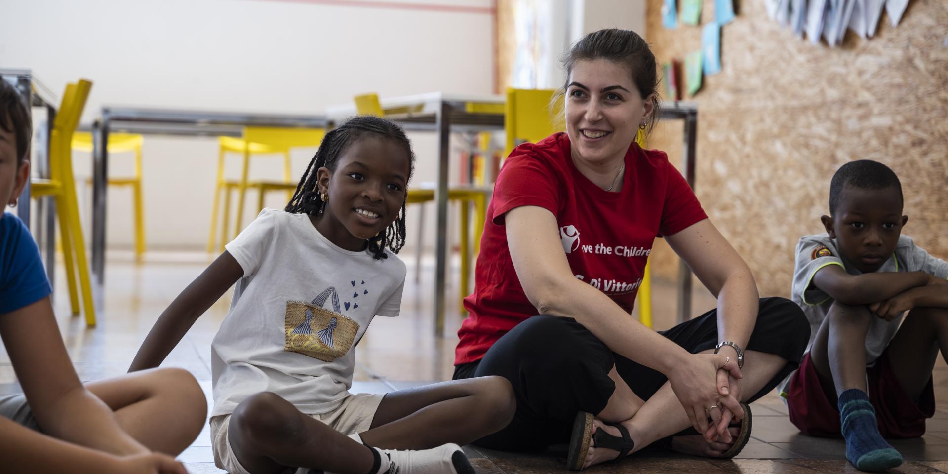 Un'operatrice con maglietta Save the Children è seduta in circolo con altri bambini e bambine durante un'attività di condivisione all'interno del Punto Luce di Prato