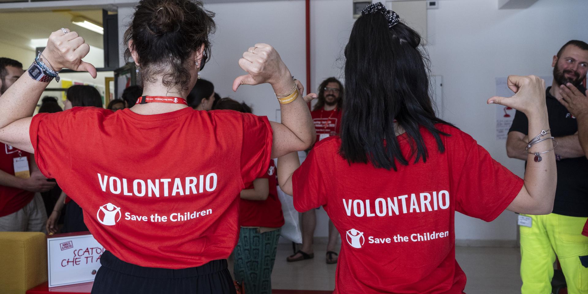 Due ragazze di spalle indicano le magliette che indossano da volontarie Save the Children durante il Meeting del Volontariato 2023.