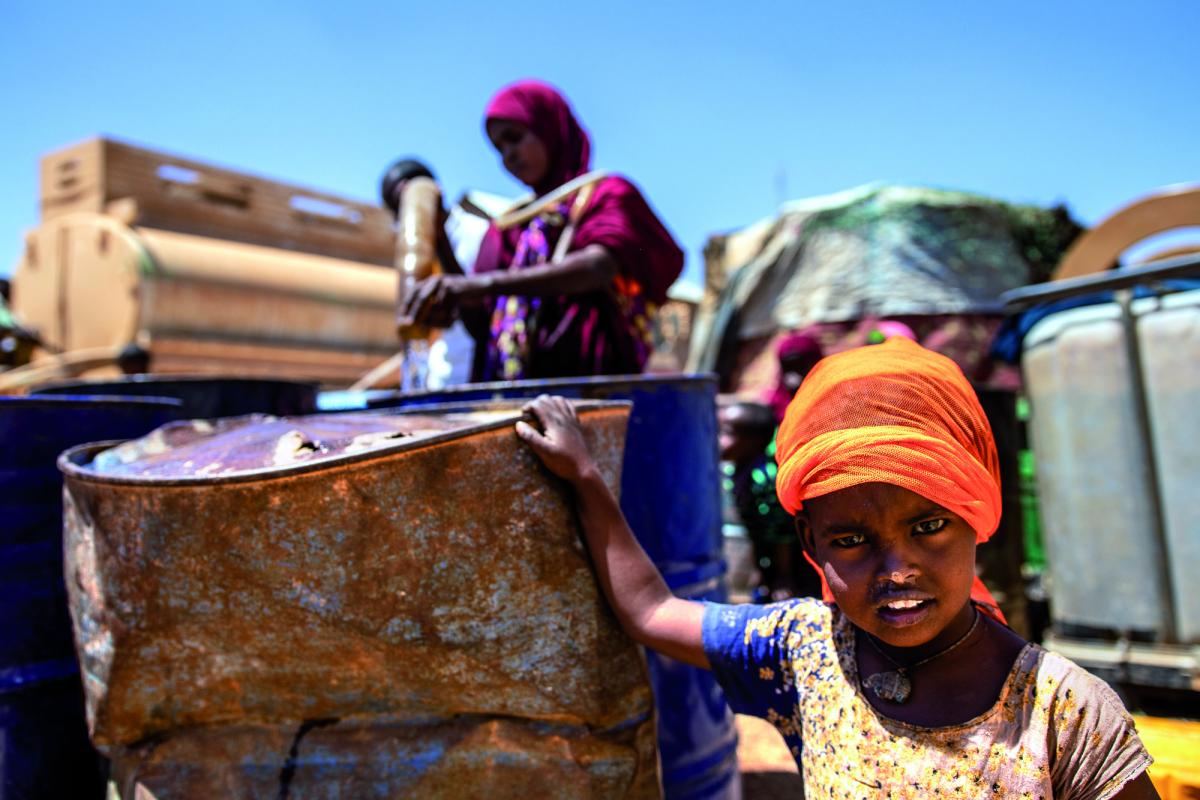 Una bimba somala fotografata accanto ad un vecchio bidone in cui sua madre sta versando dell'acqua
