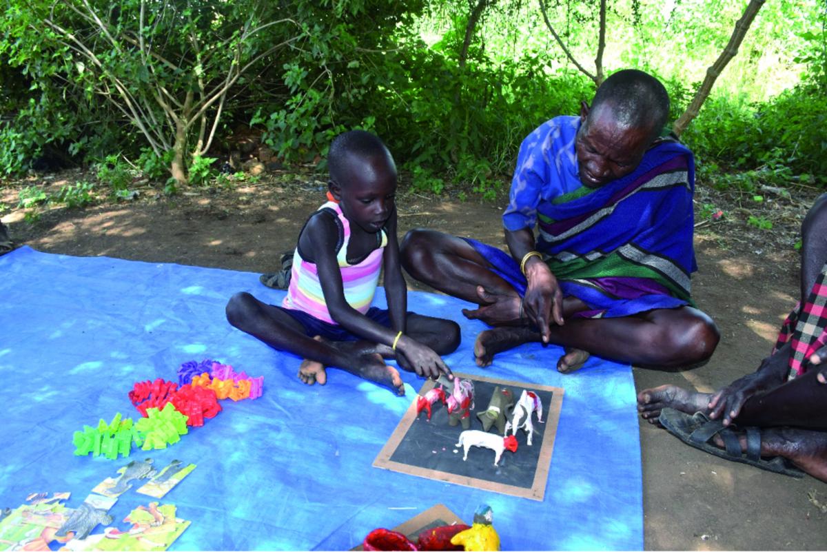 Un bambino ugandese seduto in terra mentre impara a contare con l'aiuto di una serie di pupazzetti e del suo papà formato da Save the Children