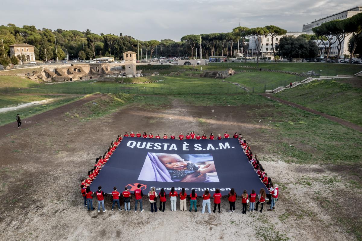 Staff e volontari di Save the Children, disposti a quadrato all'interno del Circo Massimo, tendono tra le mani un enorme telo sul quale è raffigurato una bimbina malnutrita insieme alla scritta "Questa è S.A.M.", acronimo di Severe Acute Malntrutrition, per la campagna Emergenza Fame