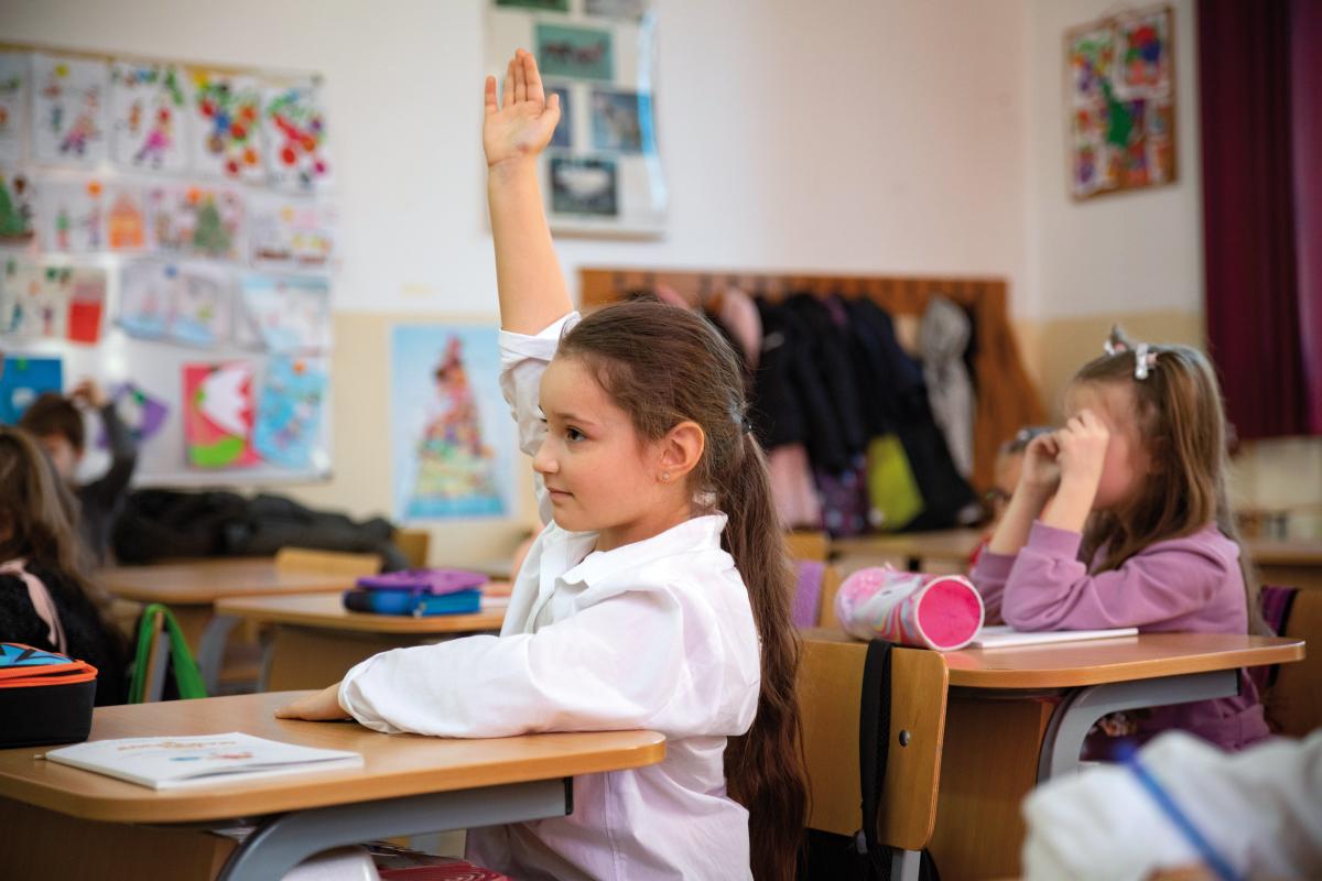 Una bambina alza la mano per intervenire a scuola. E’ seduta al banco e dietro di lei ci sono altre bambine e bambini della classe. La bambina proviene dall’Ucraina e frequenta ora una scuola in Romania.