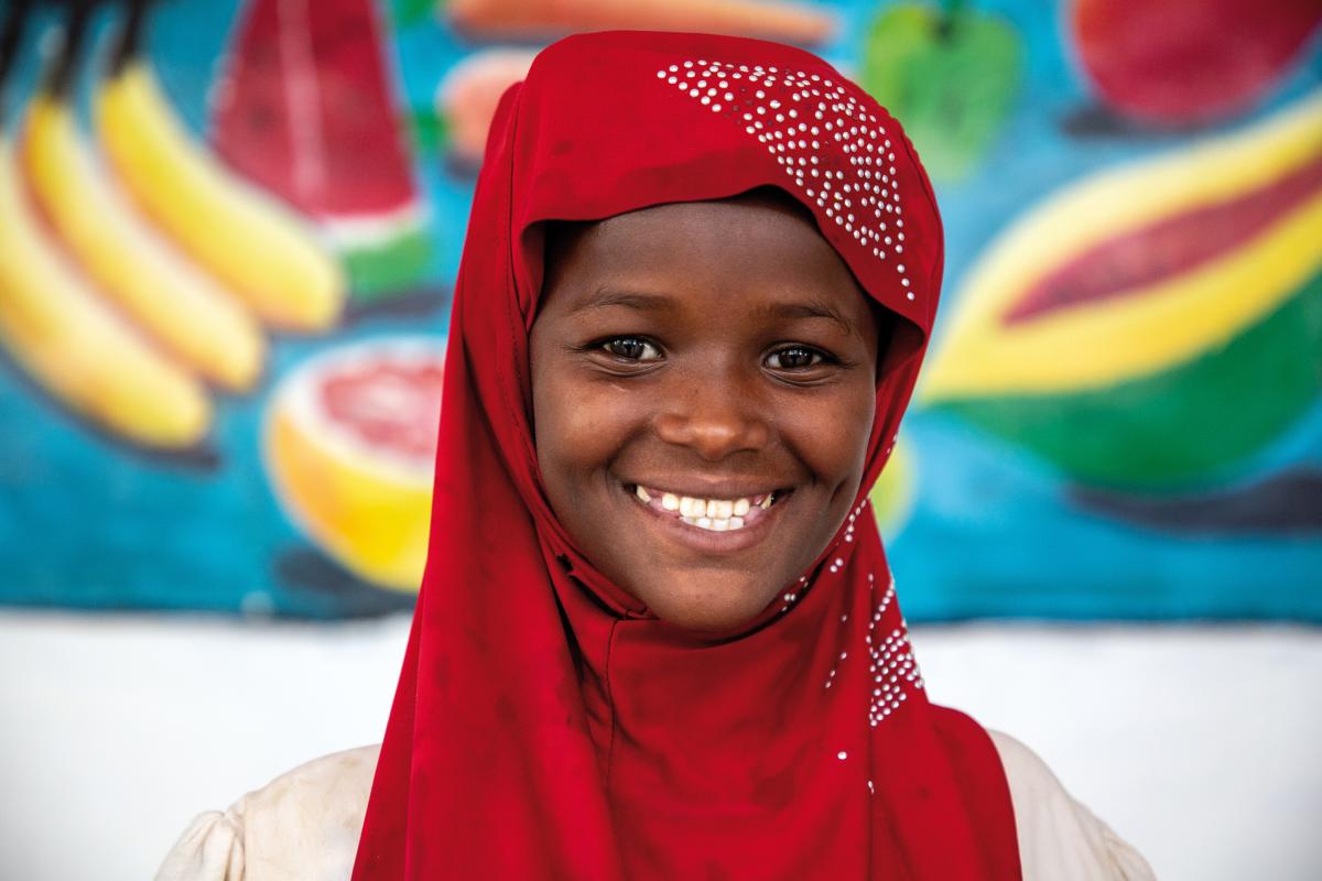 Una bambina con velo rosso sorride in camera, all’interno di un ospedale in Somalia. Sullo sfondo un dipinto di frutti molto colorato.