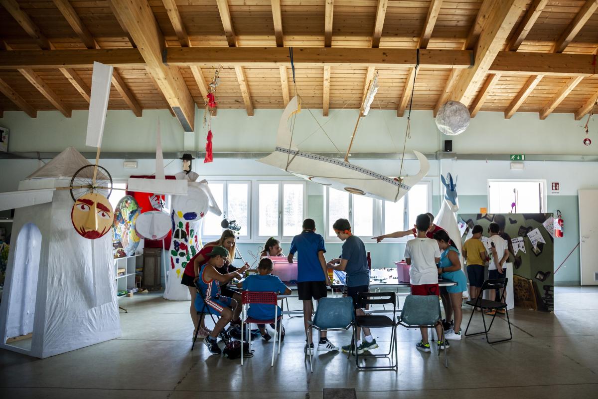 Bambine e bambini intorno ad un tavolo collaborano ad una attività ricreativa all'interno di una grande e luminosa sala del Punto Luce de L'Aquila. Dal soffitto pendono delle enormi creazioni di carta realizzate dai ragazzi del Punto Luce.