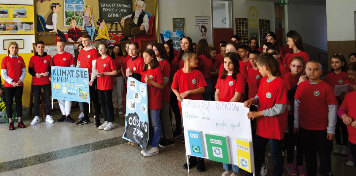 Bambini e bambine all’interno di una scuola, tutti con maglietta rossa, mostrano dei cartelloni da loro realizzati su temi ambientali come il riciclo.